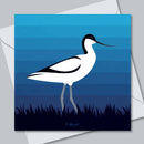 A square card featuring a stylized avocet bird in white, black, and gray against blue horizontal stripes, with dark grass silhouettes at the bottom. The print is shown slightly offset over a white envelope on a light gray background.