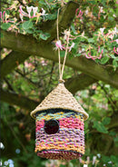 Colorful braided birdhouse hanging from a tree with flowers in the background