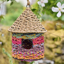 Colorful braided birdhouse hanging among greenery with a white flower.