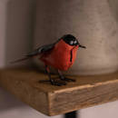 A small bird figurine with a bright red body, black head and wings, and black legs, standing on a rustic wooden shelf in front of a blurred gray ceramic vase.