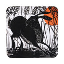 A square coaster with rounded corners featuring a black and white woodcut-style illustration of a hare among reeds, with a bright orange sun in the upper right.