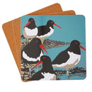 A stock of square tablemats featuring a lino print  illustration of five black-and-white oystercatcher birds with red beaks on a teal background.