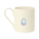 An off-white ceramic mug featuring a small, decorative speckled blue and white Easter egg illustration on the front, set against a plain white background.
