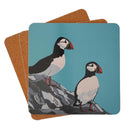 A square cork tablemat with rounded corners, featuring a printed illustration of two puffins on a teal-blue background. The coasters are stacked slightly offset against a clean white background. From Sue Welfare collection