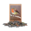 A playing card sized jigsaw puzzle box with a robin bird illustration on the front, standing upright with many puzzle pieces scattered in front on a white background.